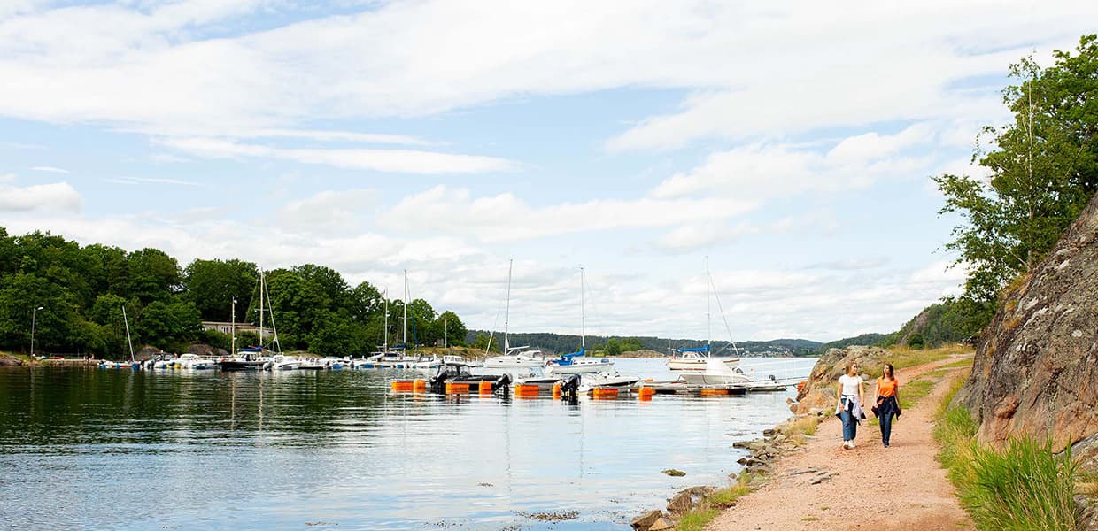 Kyststi langs småbåthavn på Veierland. To personer går på grusvei langs fjellvegg til høyre. Mange seilbåter og fritidsbåter ligger fortøyd i vannet til venstre. Blå himmel med hvite skyer.