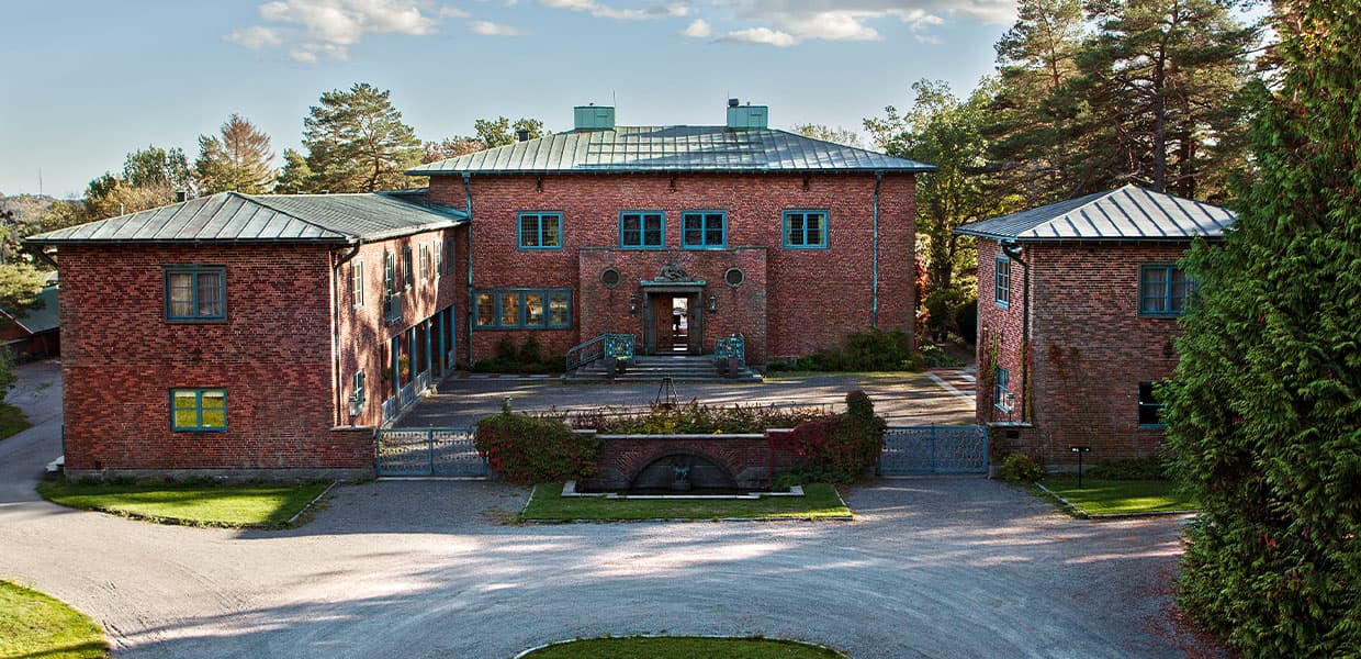 Midtåsen villa sett forfra med sin karakteristiske røde mursteinsfasade og blågrønne vinduskarmer. Bygningen har metalltak og symmetrisk utforming med to sidefløyer. En sirkulær innkjørsel og velstelt hage omgir den historiske villaen.