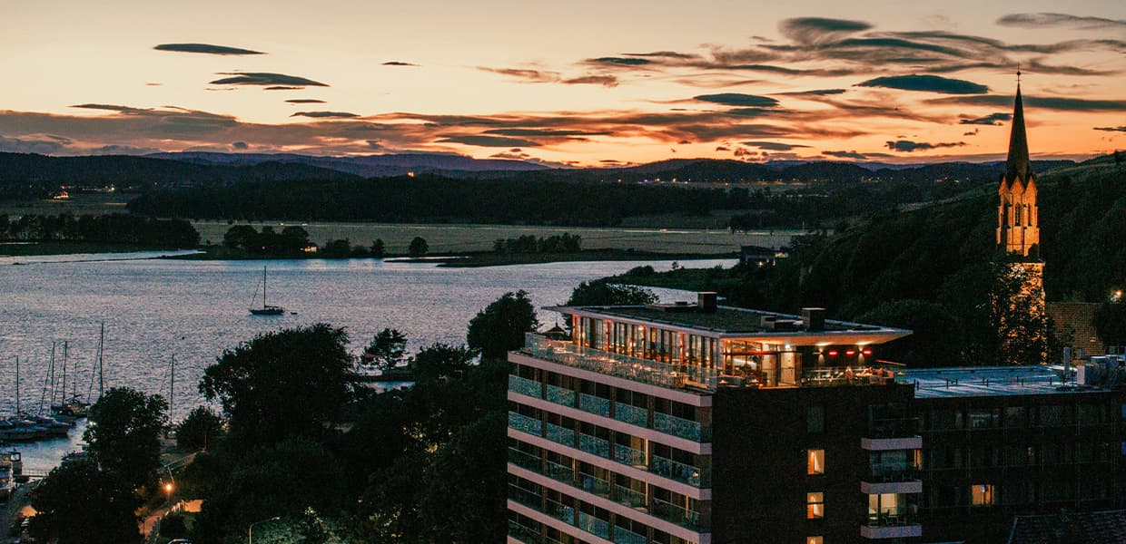 Nattutsikt over en by med vann og en kirke i bakgrunnen, en bygning med en belyst terrasse.
