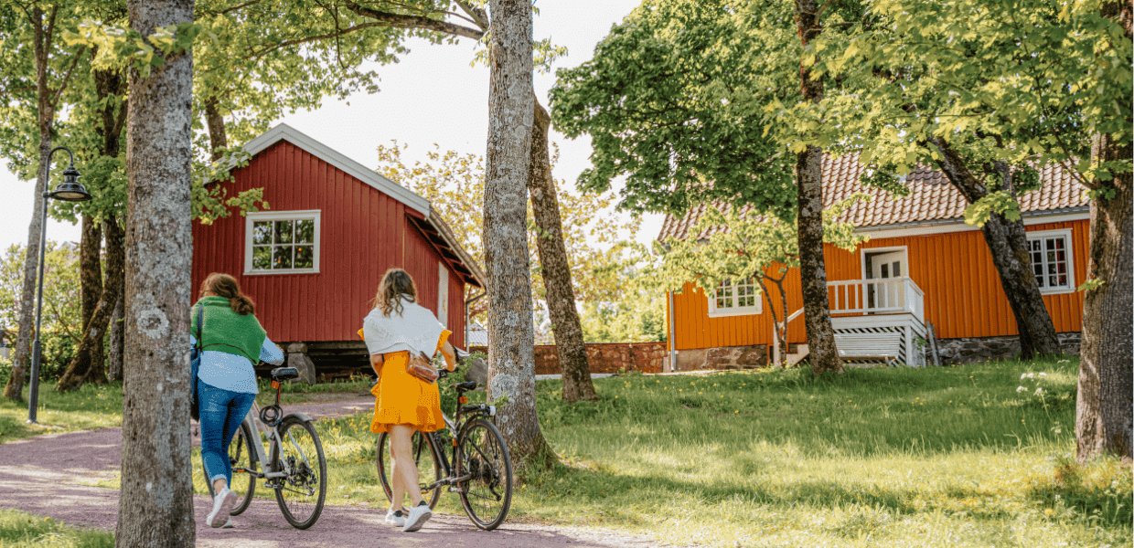to personer triller sykkel i grønn hage på grusvei foran to trehus
