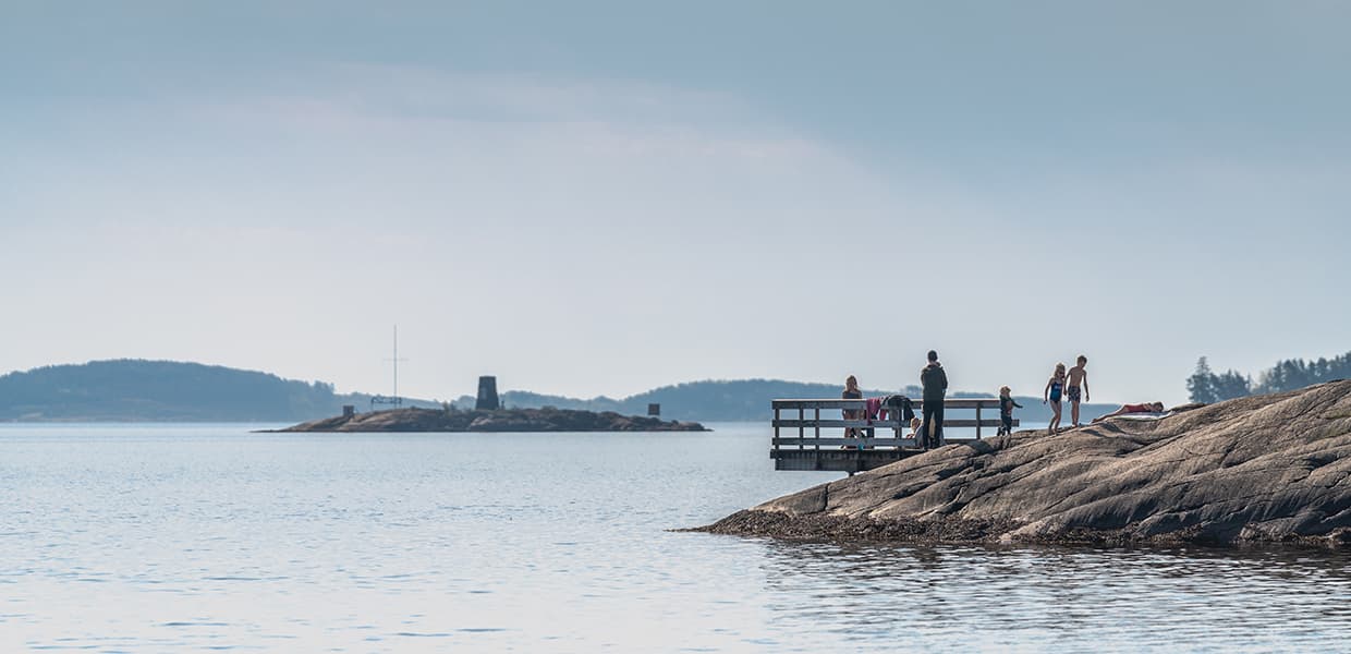 En lang trebrygge strekker seg ut fra glatte svaberg. Personer som ser ut til å ha badet står på brygga og svaberget. I bakgrunnen sees en karakteristisk fyrlykt på en holme. Vannet er stille og blått. God kontrast mellom mørke svaberg og lyst vann.