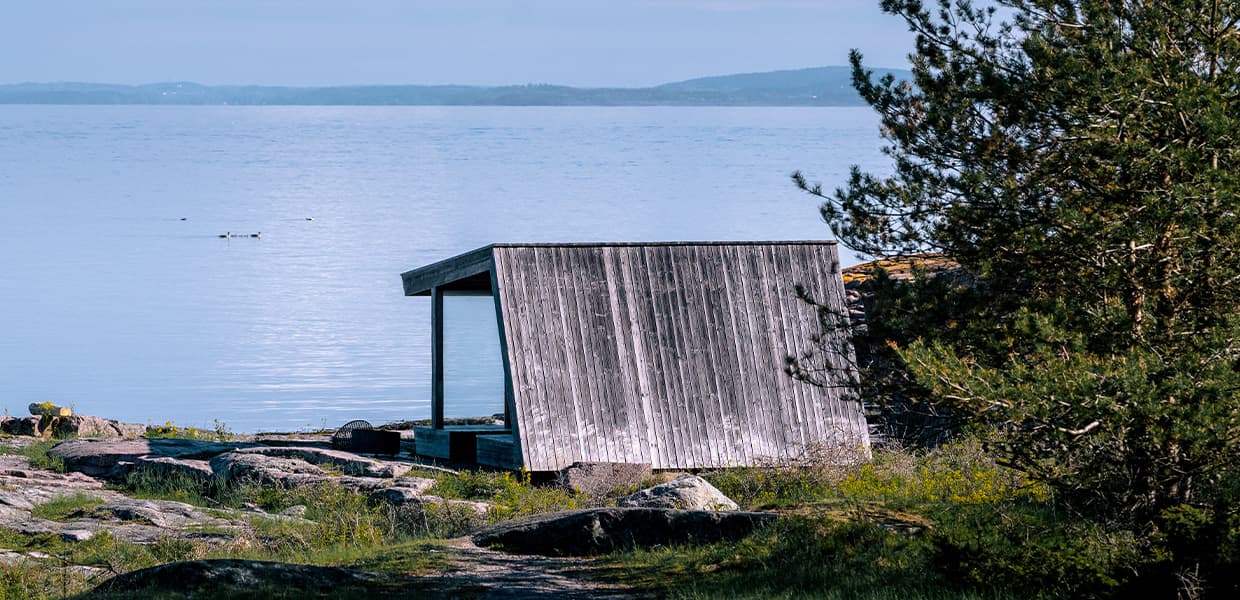 En stor trebygget gapahuk står på flate svaberg ved vannkanten. Gapahuken har grått, værslitt trepanel og skråtak. Stille, blått hav strekker seg ut foran, med noen ender som svømmer. Furutrær rammer inn bildet på høyre side.