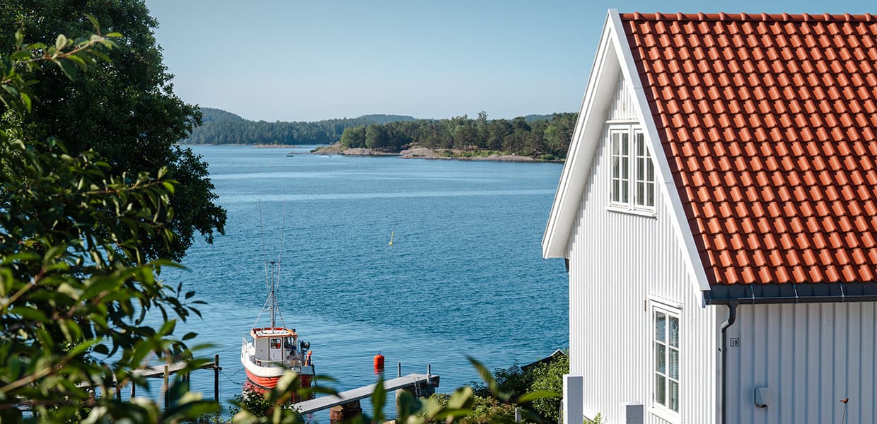 : Et stort, hvitt trehus med rødt tak står nær vannet. En rød og hvit fiskebåt ligger ved en lang trebrygge. Blått hav og grønne øyer sees i bakgrunnen. God kontrast mellom det hvite huset og omgivelsene