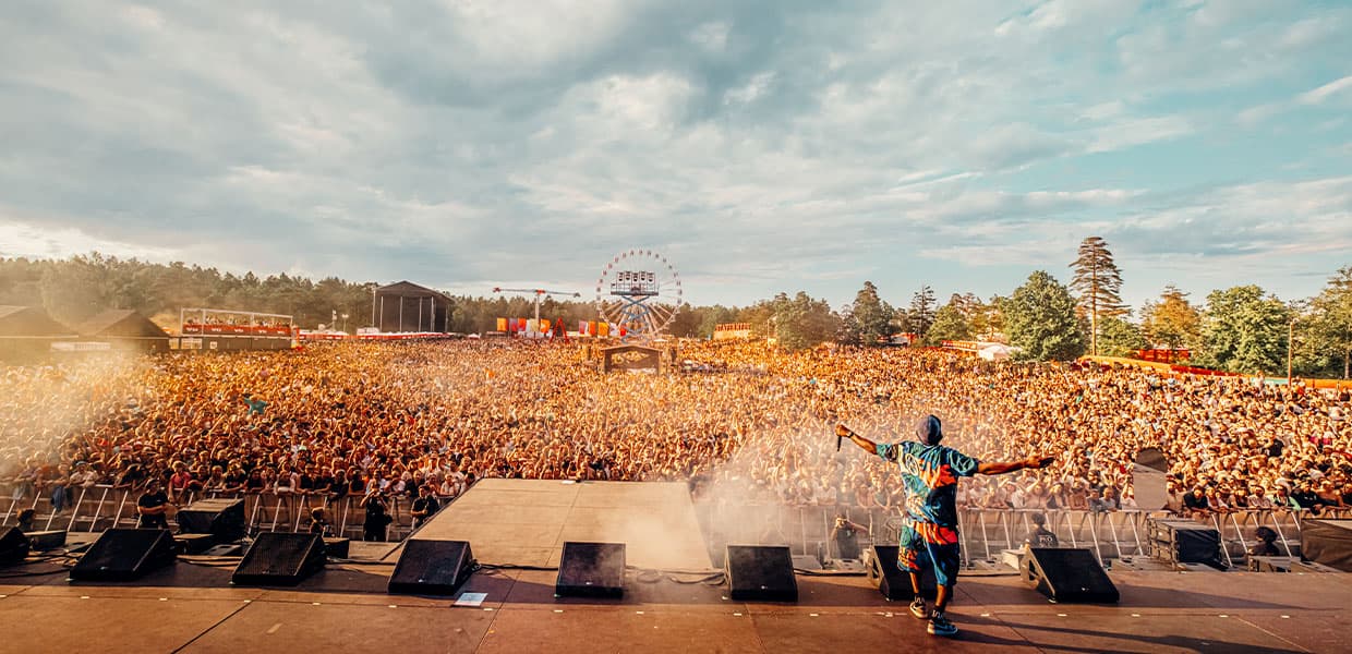 En artist i fargerik blå og oransje drakt står med armene utstrakt på scenen foran en massiv publikumsmengde på Stavernfestivalen. Tusenvis av festivaldeltakere strekker seg så langt øyet kan se under en lys himmel. I bakgrunnen sees et pariserhjul og festivalområdet med telt og trær. Scenen er badet i sollys, og det sprutes vann eller røyk foran artisten, som skaper en dramatisk effekt