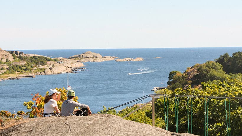 To personer med hjelm sitter på klippe med utsikt over blått hav og svaberg. Et grønt metallrekkverk markerer klatrerute og zipline.