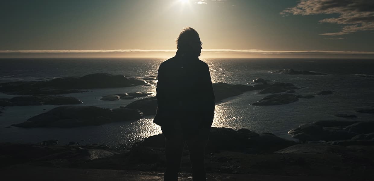 En silhuett av en person står mot en dramatisk havsolnedgang, der solen kaster en strålende glorie på himmelen og reflekterer glitrende over vannet. Scenen formidler ro og ettertanke.