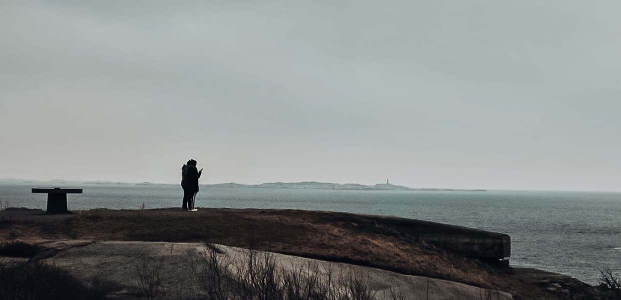 To person står på en klippeformasjon og ser utover havet. Bildet er tatt i gråtoner med disig himmel i bakgrunnen.