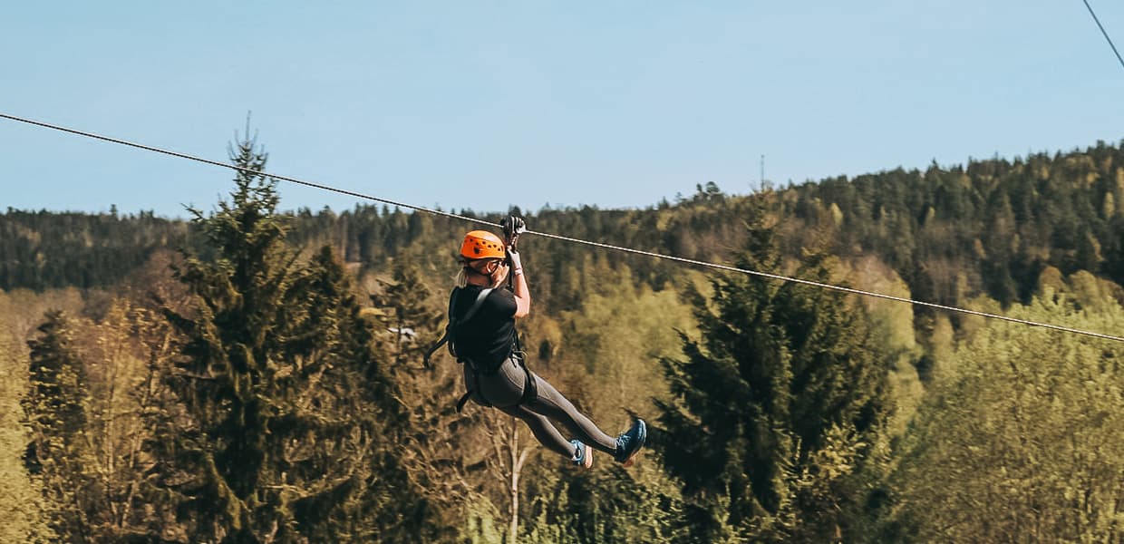 En person nyter en spennende zipline-tur over en frodig skog, ikledd hjelm og selen, og omfavner eventyret og naturens skjønnhet en solrik dag.