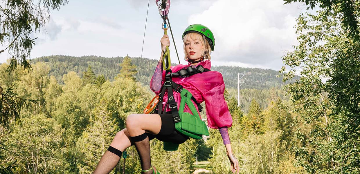 Kvinne i zipline-aktivitet svever mellom trær. Hun har på seg grønn hjelm, rosa overdel og svarte shorts, og holder seg fast i zipline-utstyret. Under henne ses en skog av løvtrær i høstfarger, og hun er fotografert mot en lys himmel.