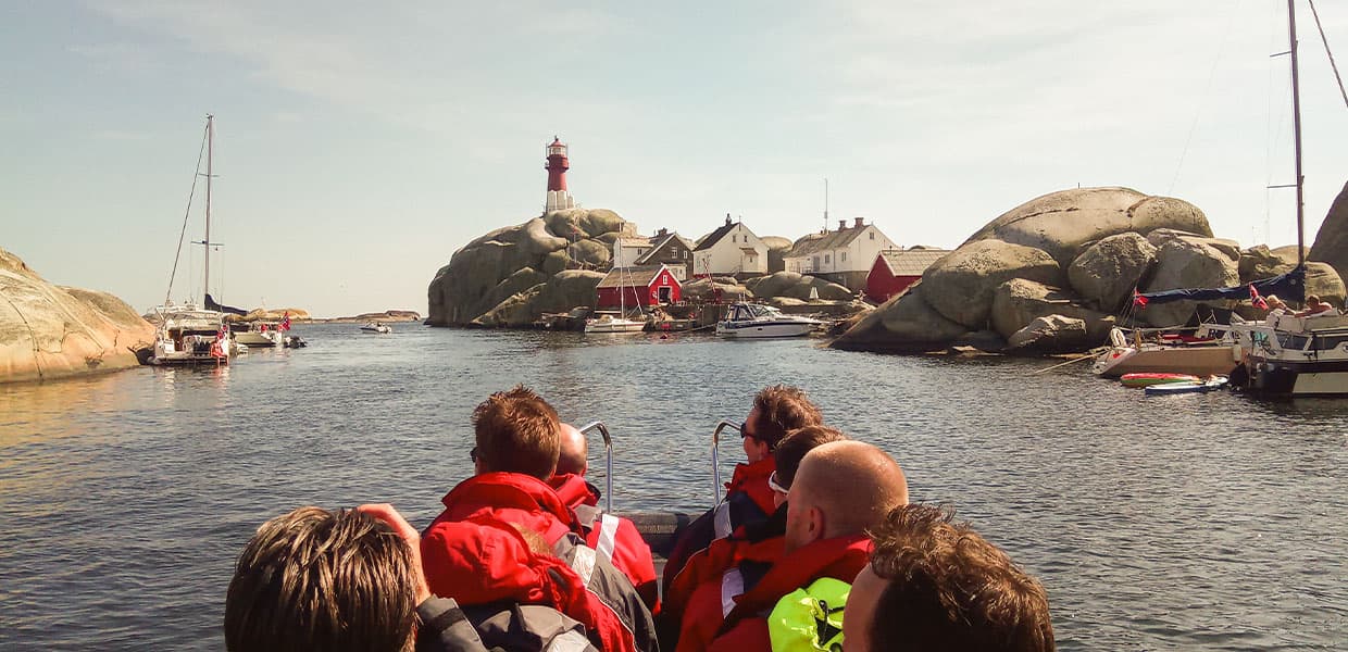 Passasjerer med røde redningsvester i forgrunnen ser mot det røde fyrtårnet som rager over hvite og røde hus bygget mellom store svaberg. Flere fritidsbåter ligger fortøyd i den beskyttede havnen. Sterk kontrast mellom det blå vannet, det røde fyret og de lyse svabergene.