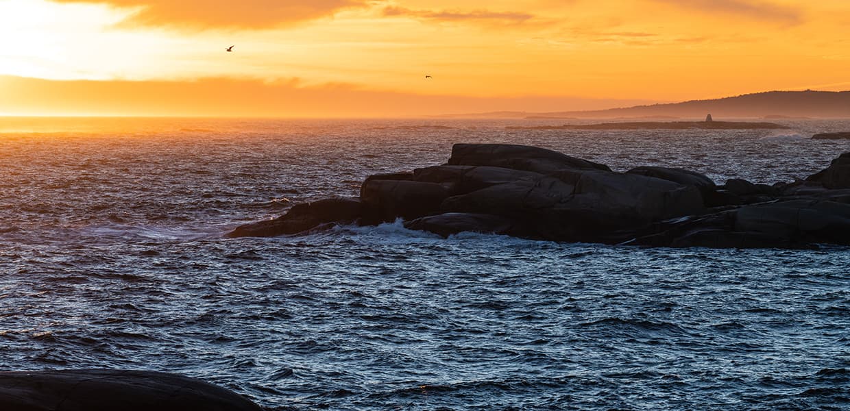 Solnedgang over Verdens Ende på Tjøme. Det historiske utkikkstårnet og bygninger på en liten odde er silhuettert mot en dramatisk himmel i rosa og oransje farger. Det stille vannet reflekterer himmelens farger.