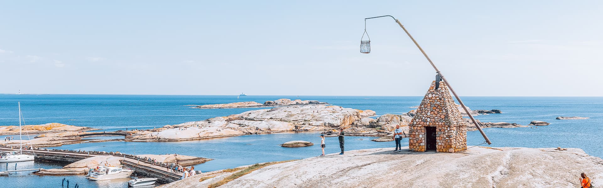 Et ikonisk steinhus med pyramideformet tak står på klippeformasjoner ved kysten. En høy mast med en kurv strekker seg over taket. Det klare, blå havet bryter mot de steinete øyene i bakgrunnen.