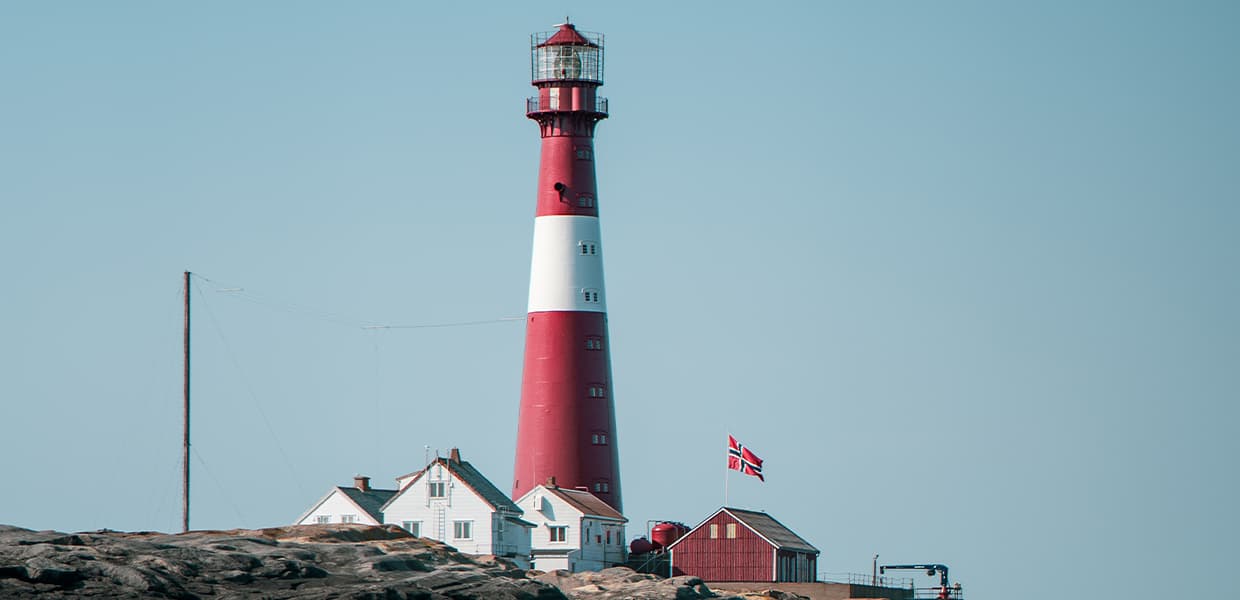 Det rød-hvite fyrtårnet står på en steinete grunn og overseer havet. Det norske flagget henger ved siden av, og bygningene har en hvit og rød stil som kontrasterer med den blå himmelen.