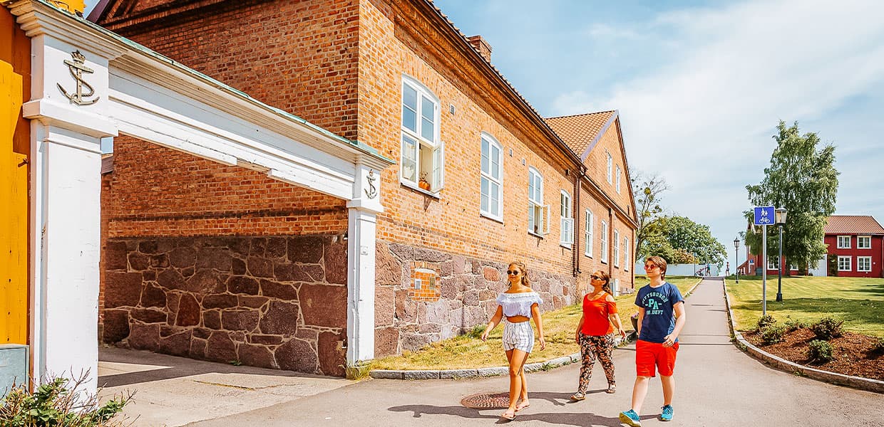 Tre personer går langs en solfylt gate på Karljohansvern i Horten, omgitt av historiske murbygninger og grønne områder. De passerer en hvit port med marineemblemer, en påminnelse om områdets maritime historie.