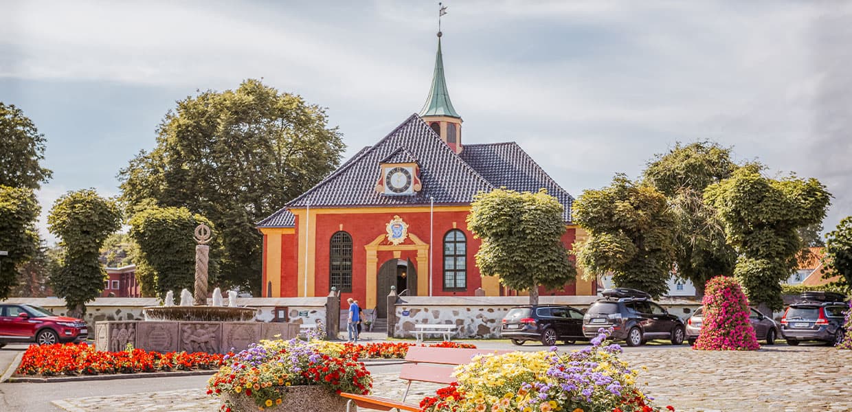 En gul og rød kirke med grønt tårn omgitt av trær og blomsterbed.