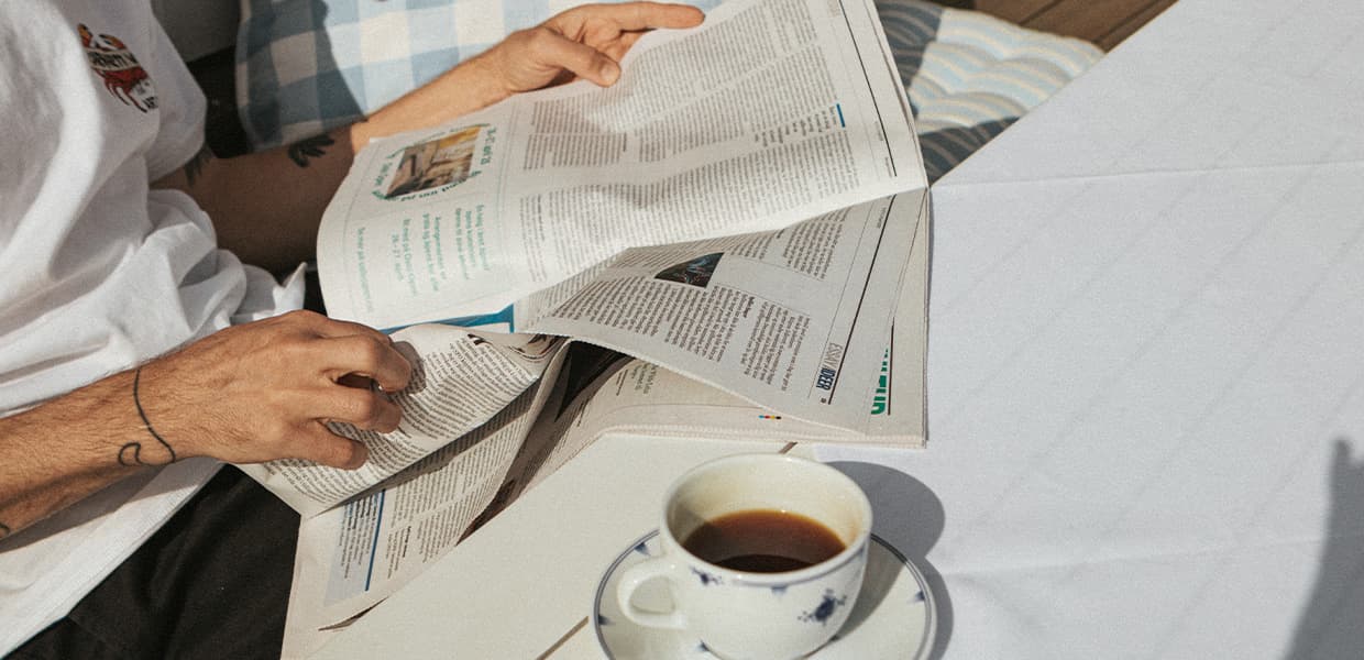 person leser avis utendørs i sola med en kaffekopp ved siden av