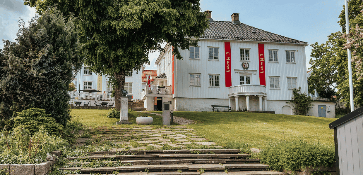 Hvitmalt bygning med røde bannere på fasaden, sett fra en steintrapp med gressplen og trær i forgrunnen.